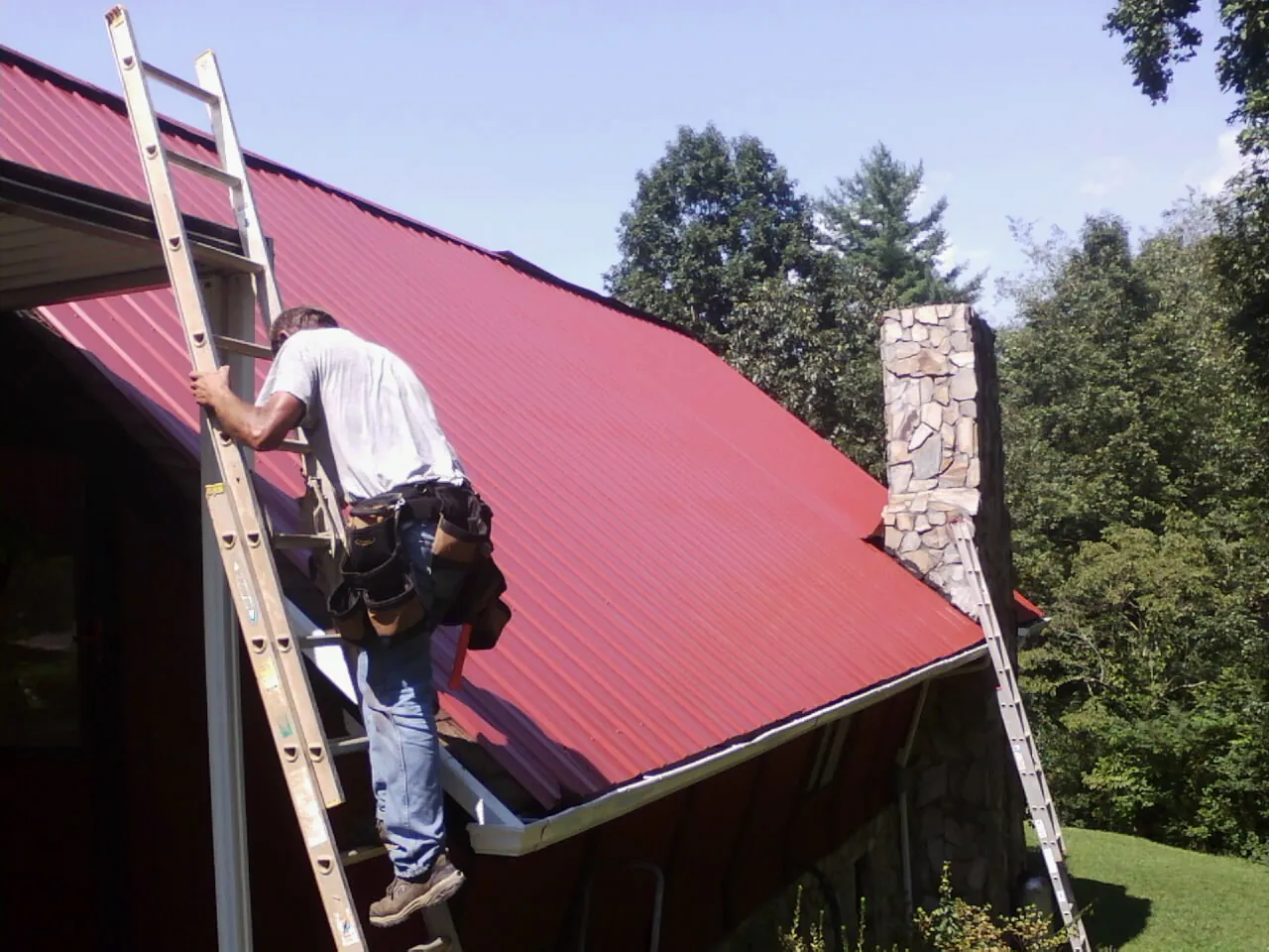 Roofer on a ladder beside a red metal roof and stone chimney — Soffit & Fascia Repair by Roofing Brothers in Walbridge